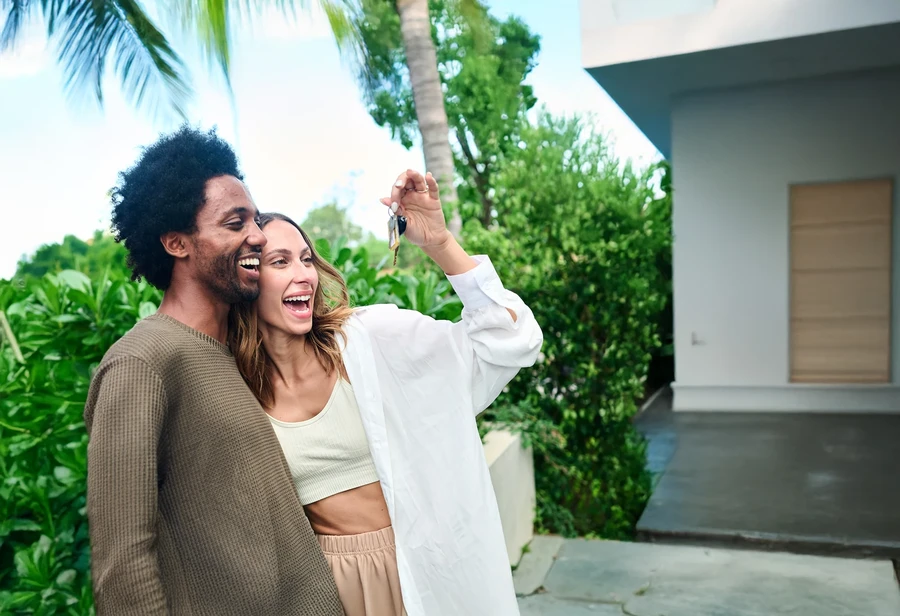 Happy couple holding up a house key outside a modern Florida home with tropical plants